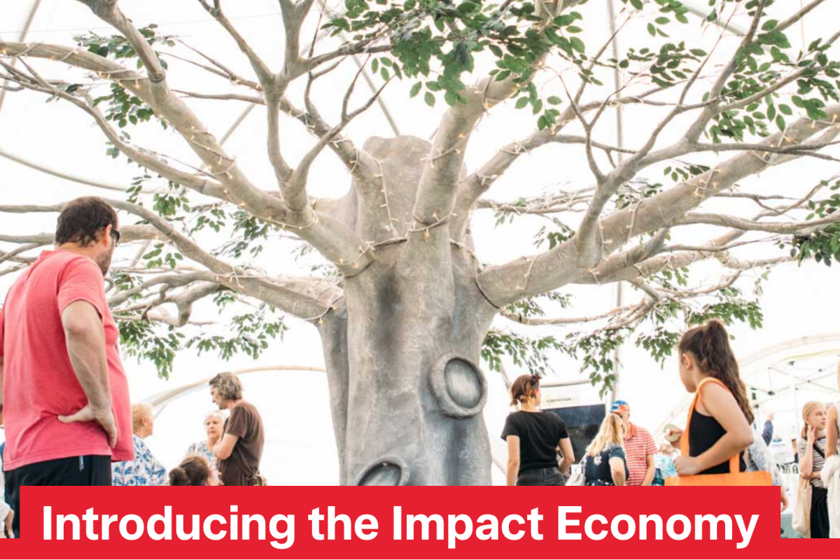 Group of people gathered around a large artificial tree with green foliage under a bright structure, featuring the text 'Introducing the Impact Economy' in bold red at the bottom.